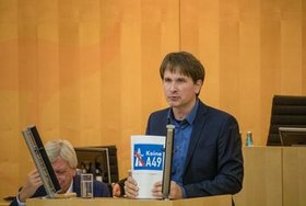 Jan Schalauske steht am hellgrauen Redner:innenpult im Hessischen Landtag mit einem Bericht zur A49 in der Hand.