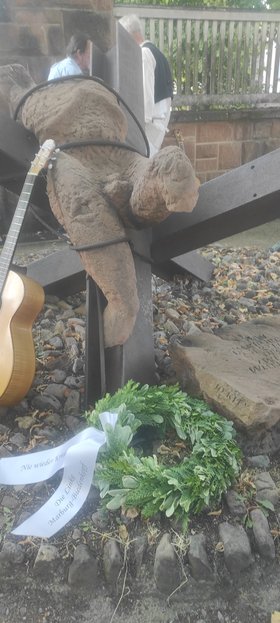 Ein Blätterkranz mit Widmungen liegt vor einem flachen Denkmal auf dem Boden; im Hintergrund eine angelehnte Gitarre