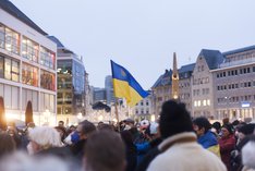 Demonstration in einer Innenstadt für Solidarität mit der Ukraine. Man sieht eine undeutliche, aber große Menschenmenge und in der Mitte des Bildes eine Person, die eine ukrainische Flagge hochhält.