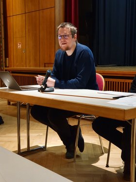 Ein junger Mann sitzt an einem Tisch und diskutiert mit Publikum.