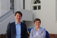 Die Spitzenkandidat*innen der LINKEN, Jan Schalauske und Elisabeth Kula, stehen vor einer weißen Wand lächelnd in der Sonne.