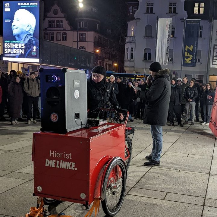 Ein Mann in dunkler Kleidung steht neben einem Aufbau mit Lautsprecher und hält eine Rede