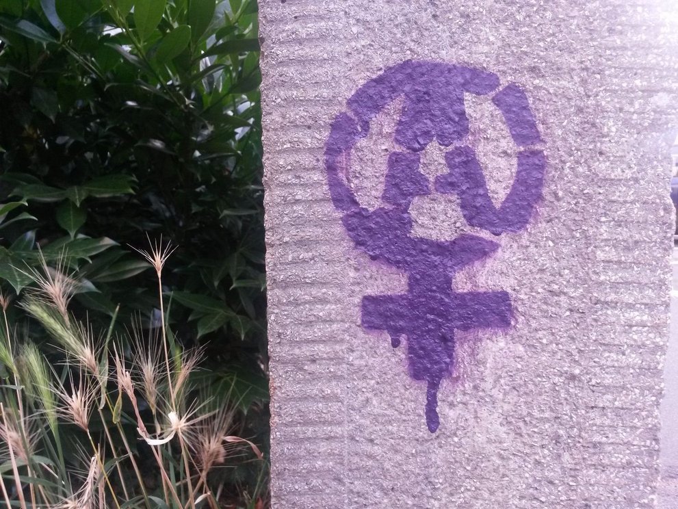 Das Symbol für Frau* mit einem A in der Mitte in lila auf eine grau-rosa Hauswand gesprayt. Links davon Pflanzen.
