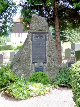 In einem grünen Park unter einem großen Baum erinnert ein Stein mit einer Kupferplakette an die Morde von Mechterstädt, begangen von Marburger Studenten.
