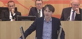 Jan Schalauske steht am hellgrauen Redner:innenpult im Hessischen Landtag mit dem Präsidium im Hintergrund