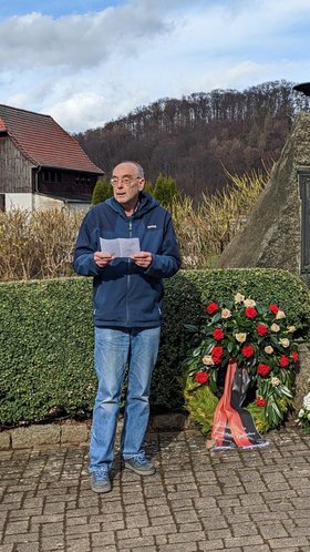 Ein Mann hält neben einem Gedenkstein in der Frühlingssonne eine Rede.